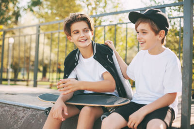 Two cute teenagers sit in a skatepark, relax after skateboarding and chat. 