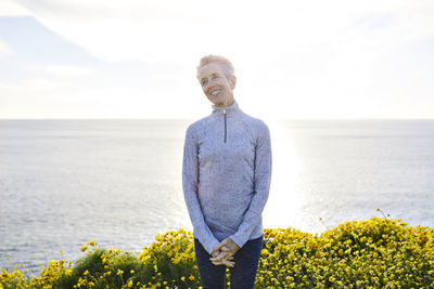 Smiling senior woman standing by flowering plants by sea against bright sky