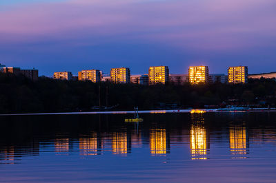 Illuminated buildings by lake against sky at dusk