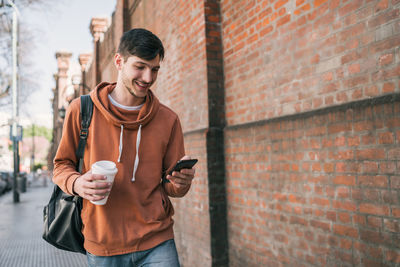 Young man listening music over mobile phone while holding coffee cup against brick wall