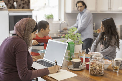 Muslim woman working on laptop with family having breakfast in kitchen