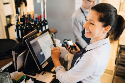 Smiling woman using computer while holding credit card reader in restaurant