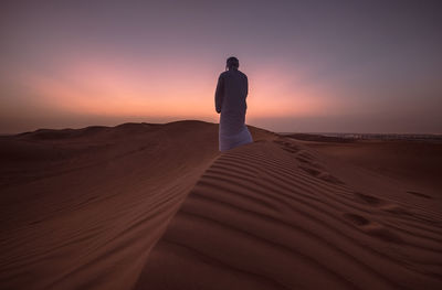 Rear view of man standing on sand dune at desert against sky during sunset