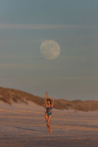 Slim female in swimsuit standing with raised arms on sandy beach in evening under blue sunset sky with moon