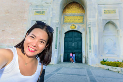 Portrait of smiling young woman taking selfie against built structure