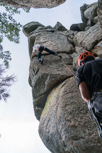 From below climber in helmet holding rope and insuring partner crawling on sheer mountain