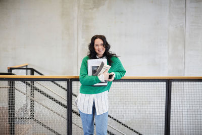 Portrait of smiling student with books and mobile phone standing at railing against wall in university