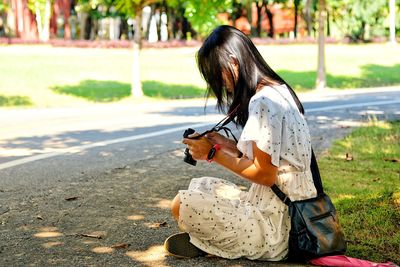 Side view of woman using camera while sitting by road