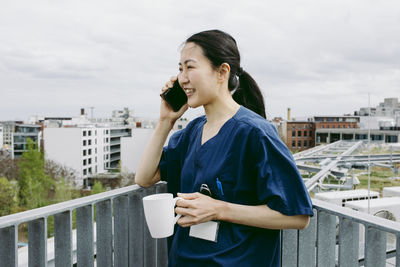 Smiling mid adult female nurse holding coffee cup while talking on smart phone