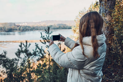 Young woman taking photos of landscape with her smartphone during trip on autumn sunny day