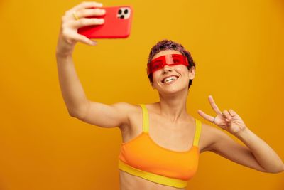 Portrait of young woman taking selfie against yellow background