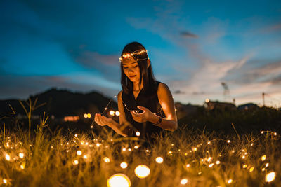 Woman with illuminated string light sitting on grass during sunset