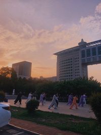 People sitting by buildings against sky during sunset