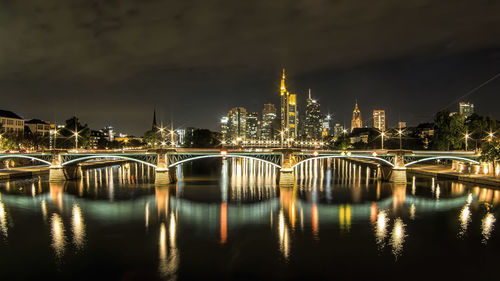 Illuminated bridge over river by buildings against sky at night