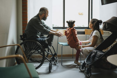 Mother and daughter looking through window by man in wheelchair at hospital