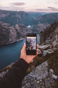 Hand holding smartphone with image of scene in background on it at norwegian fjords