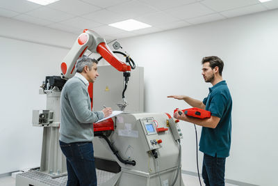 Male businessman with control discussing with mature engineer at machinery in factory