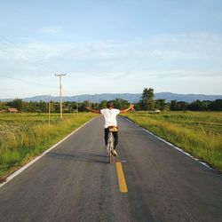 Rear view of man cycling with arms outstretched on road against sky