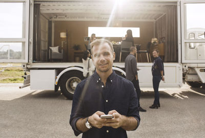 Portrait of businessman holding mobile phone with colleagues and portable office truck in background