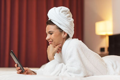 Side view of woman wearing towel while using mobile phone on bed at home