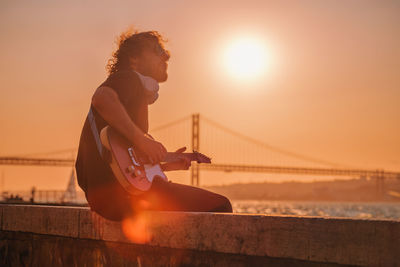 Side view of young man playing guitar against sky during sunset