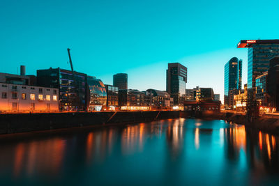 Illuminated buildings by river against blue sky at dusk