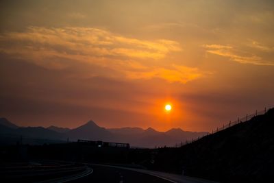 Road by silhouette mountains against orange sky