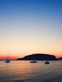 Boats at sea by silhouette mountain against clear sky during sunset