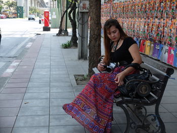 Woman holding camera while sitting on bench on sidewalk