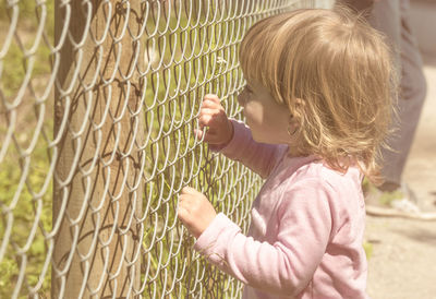 Side view of cute girl looking through chainlink fence during sunny day