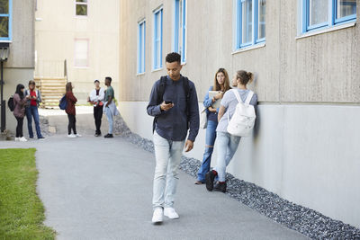 Young man using mobile phone while walking at campus with students standing in background