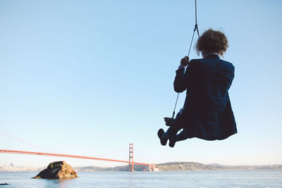 Rear view of young woman swinging at beach against clear sky