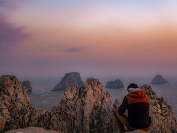 Rear view of people sitting on rock against sky during sunset