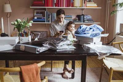 Working mother using smart phone while sitting with baby boy at home office