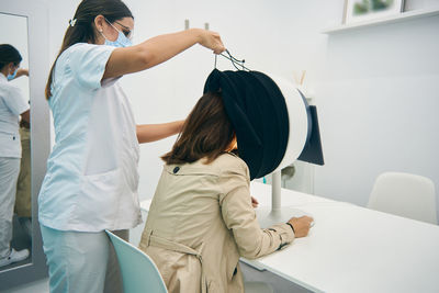 Side view of female cosmetologist using modern facial diagnostics machine for analyzing skin of client in beauty clinic