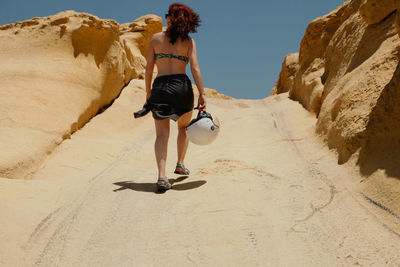 Rear view of women holding helmet while walking on sand