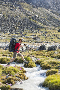 Backpacker steps across river in alpine meadow.