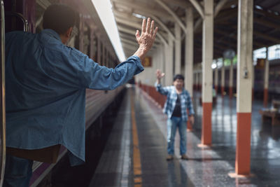 Rear view of man waving hand to friend while standing in train at railroad station platform