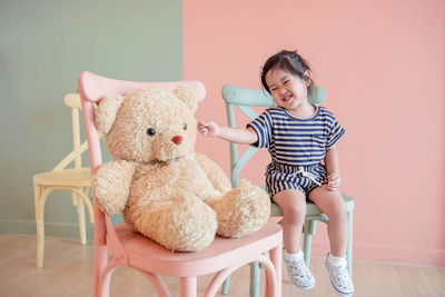 Full length of cute girl sitting with teddy bear on chairs at home