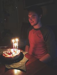 Young man looking at burning candles on birthday cake in dark