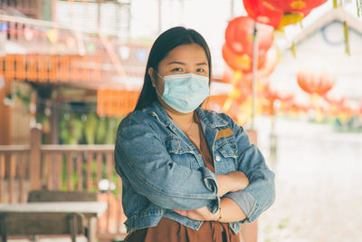 An asian woman in casual clothes wearing a mask and crossing her arms looking at a camera