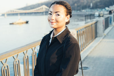 Portrait of smiling young woman standing by railing and sea
