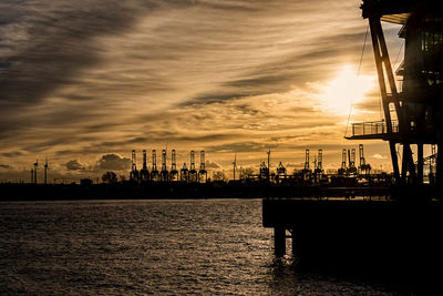 Silhouette cranes at harbor against sky during sunset