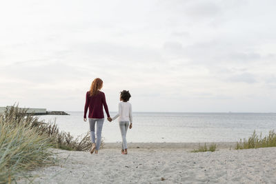 Rear view of mother and daughter holding hands while walking on sand at beach against sky