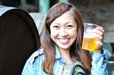 Smiling young woman holding beer glass against barrels on sunny day