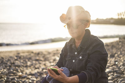 Senior woman using mobile phone while sitting at beach during sunset