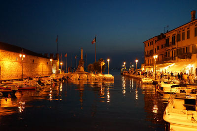 Sailboats moored in illuminated canal by buildings against sky at night