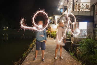 Friends making love text with sparkler while standing by lake against sky at night