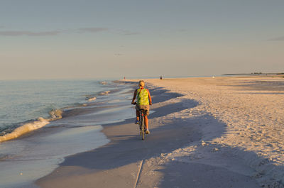 Full length rear view of person riding bicycle at beach against sky on sunny day