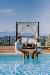 Couple sitting by swimming pool against sky